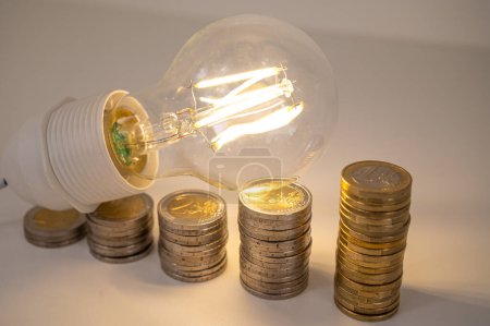 Light bulb lit, above stacks of coins. Increase in electricity tariffs, energy dependence, energy supplies.