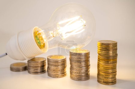 Light bulb lit, above stacks of coins. Increase in electricity tariffs, energy dependence, energy supplies.