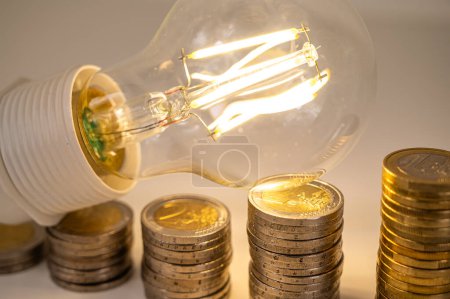 Ampoule allumée, au-dessus des piles de pièces. Augmentation des tarifs d'électricité, dépendance énergétique, approvisionnement énergétique.