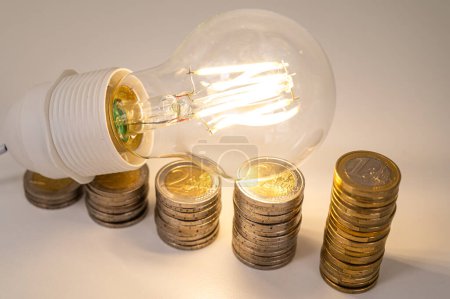 Glühbirne angezündet, über Stapeln von Münzen. Erhöhung der Strompreise, Energieabhängigkeit, Energieversorgung.