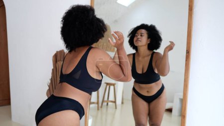 Foto de Multicultural plus size woman in lingerie looking at her body in the mirror. Black woman struggles with obesity and overweight, wants to lose weight and be slim Woman body positive without criticism - Imagen libre de derechos