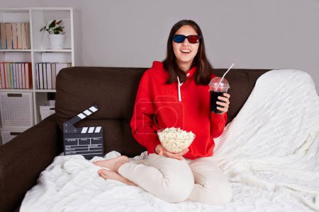 Photo for A young smiling girl with 3d glasses is watching a movie at home. A teenager holds a glass with a drink and popcorn in his hands. - Royalty Free Image