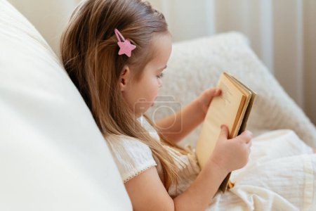 Foto de Pequeña chica rubia linda leyendo libro sentado en un sofá. Lectura infantil, ensueño y desarrollo de la imaginación. Día Internacional del Libro Infantil. A los niños les encanta leer. Día Mundial del Libro. - Imagen libre de derechos