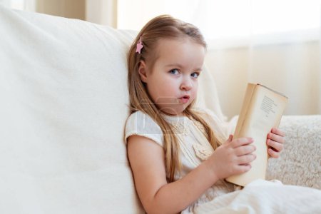 Photo for Little cute blond girl reading book siting on a sofa. Child reading, dreaming and imagination development. International Children's Book Day. Kids Love to reading. World Book Day. - Royalty Free Image