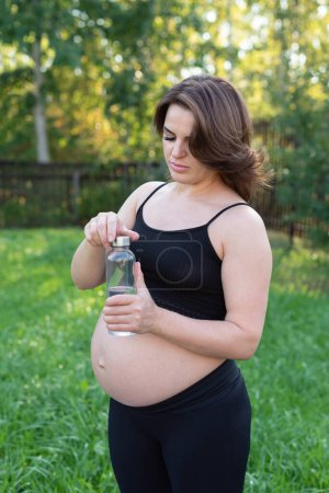 Photo for Pregnant woman in sportswear drinking a bottle of water after doing yoga outdoors, taking break in yoga practice, fitness and meditating. Motherhood, mental health and pregnancy concept. - Royalty Free Image