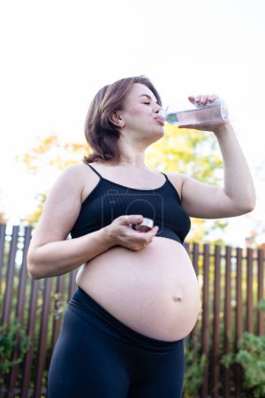 Foto de Mujer embarazada en ropa deportiva bebiendo una botella de agua después de hacer yoga al aire libre, tomando un descanso en la práctica de yoga, fitness y meditación. Maternidad, salud mental y concepto de embarazo. - Imagen libre de derechos