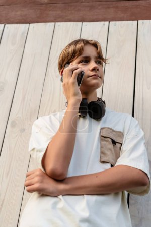 Foto de Adolescente niño usando auriculares y hablando por teléfono hablando de una conversación al aire libre, niño utilizar el teléfono celular móvil. Conversaciones al aire libre, adolescente hablando en el teléfono inteligente. - Imagen libre de derechos