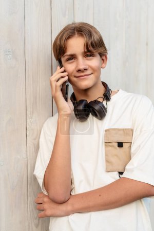Photo for Teen boy wearing headphones and talking on the phone talking a conversation outdoor, kid use mobile cell phone. Outdoor conversations, teenager speaking on smartphone. - Royalty Free Image