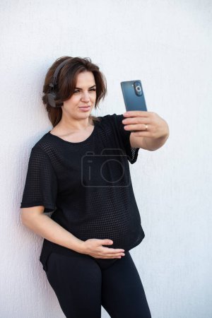 Foto de Mujer joven embarazada tomando selfie con teléfono móvil o haciendo videollamadas al aire libre. Maternidad, salud mental y concepto de embarazo. - Imagen libre de derechos