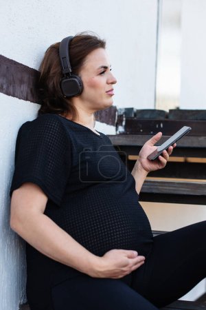 Photo for Pregnant woman with smartphone listening music in headphones outdoor on white background, spending free time relaxing. Technology, motherhood, mental health, stress relief, mediation and pregnancy. - Royalty Free Image