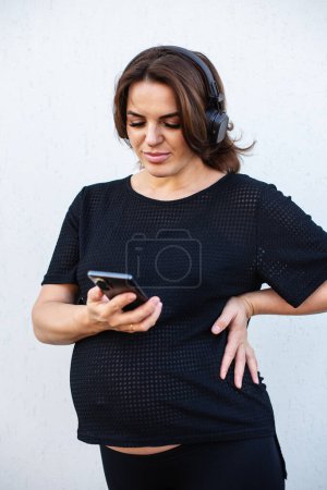 Photo for Pregnant woman with smartphone and headphones outdoor on white background. Motherhood, mental health and pregnancy concept. Social media, internet business, chatting, conversation on mobile phone. - Royalty Free Image