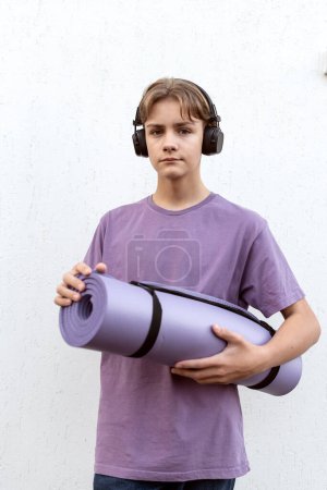 Photo for Teen boy with headphones holding yoga mat outdoor on white background. Child spending free time relaxing, listening music. Technology, teen hobby, lifestyle and people concept. - Royalty Free Image