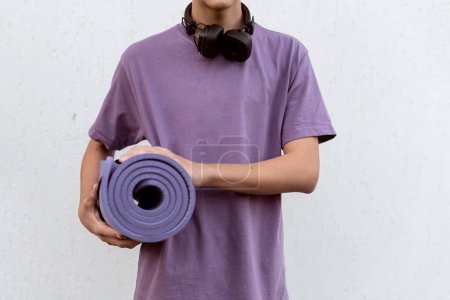 Photo for Teen boy with headphones holding yoga mat outdoor on white background. Child spending free time relaxing, sport, yoga, walking, listening music. Teen hobby, lifestyle and people concept - Royalty Free Image