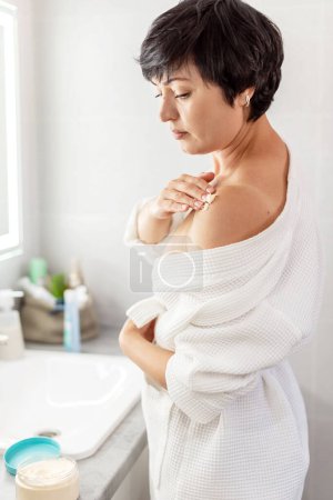 Photo for Beauty routine and body care. Cream, skincare and ethnic middle aged woman applied cosmetic moisturizing cream lotion on her body in bathroom. Beauty treatment at home - Royalty Free Image