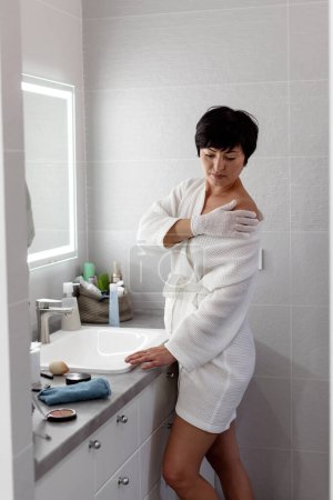 Foto de Mujer de mediana edad étnica en blanco con guante de baño exfoliante en el baño, hace un guante de masaje de pies para el cuerpo y los pies. Cosmetología, exfoliación, cosmética de spa de belleza masaje de piel y cuerpo. - Imagen libre de derechos