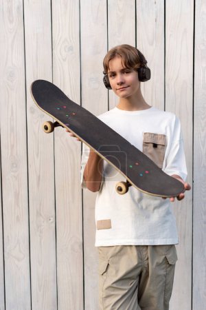 Photo for Teen boy in headphones holding skateboard outdoor on wooden white background. Child spending free time relaxing, walking, listen music. Technology, teen hobby, active lifestyle and people concept. - Royalty Free Image