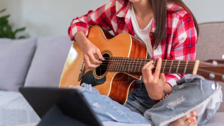 Concept of relaxation with music, Young woman plays acoustic guitar while learning music on tablet.