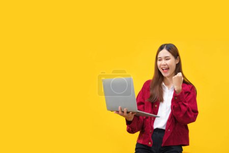 Téléchargez les photos : Les femmes tiennent un ordinateur portable et lèvent le bras pour célébrer avec un espace vide sur fond jaune isolé. - en image libre de droit