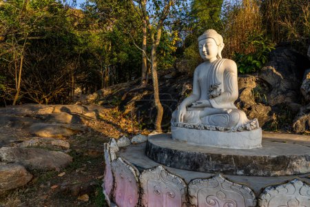 Foto de White meditating Buddha statue sits on the rock pile among forest trees in the afternoon time at India. Great religious architecture. Hight quality photo - Imagen libre de derechos