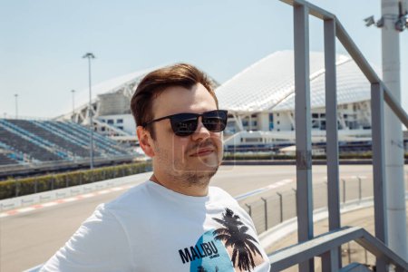 Photo for Young caucasian 34 years old man with bristle in black sunglasses over empty race car track, motorcycle racetrack. Headshot man lifestyle portrait. - Royalty Free Image