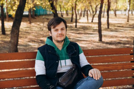 Photo for Looking at camera young caucasian unshaven man 34 years old man in sweatshirt and warm vest sits on bench in autumn park. Outdoor park, sunny day, waist up lifestyle portrait. Waiting concept - Royalty Free Image