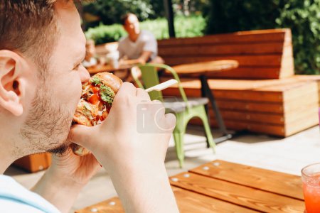 Young hungry caucasian man eating hamburger or beef burger with vegetables and sauce in outdoor street cafe. Having brunch on sunny day. Close-up, fastfood, portrait.