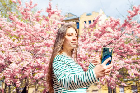 Portrait macht Selfie weiblich auf Smartphone auf Kirschblütensakurabaum. Junge lächelnde Kaukasierin macht Selfie-Porträt mit Handy. Sommer Frühling Taille nach oben Lifestyle-Porträt.