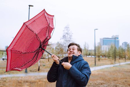 Schockierter junger kaukasischer europäischer weißer Mann mit kaputtem Regenschirm im Stadtpark, starkem Sturmwind, Regen und schlechtem Wetter. Wind riss Schirm auf. Herbst Taille nach oben Lifestyle-Porträt.