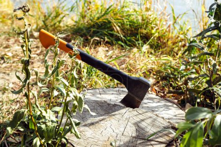 Photo for Universal orange red black modern axe with Shock absorbing fibreglass handle on dry stump log on lake coast in grass. Hiking and picnic. - Royalty Free Image