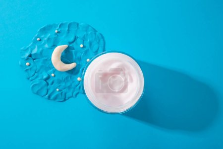 Photo for Cosmetology product for night facial care. Moisturizer cream in open glass jar and plasticine modeling clay moon and stars on blue background. Skincare, moisturizing beauty cream, unbranded moisturize - Royalty Free Image