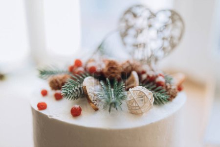 Foto de White holiday white cake covered with chocolate coins, snowball and spruce tree branches, pinecones and wooden heart. On the cake, the number 2021 is put out in red berries. The concept of a New year. - Imagen libre de derechos