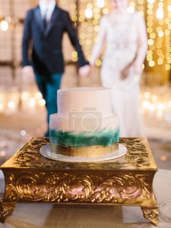 Foto de Un pastel de boda de dos niveles con decoración de oro y verde se encuentra en un soporte de oro. En el fondo, los recién casados se toman de la mano. - Imagen libre de derechos