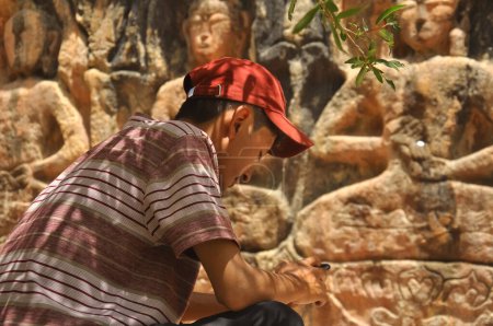 Téléchargez les photos : Rear view of a ladakhi young guy with wearing cap and using phone while sitting in front of Gyalwa Ringna 5 Dhyani Buddha rock statue which is located in Zanskar, Ladakh, India - en image libre de droit