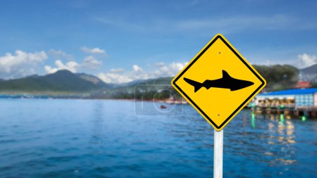 Photo for Graphic of shark on yellow sign on blurred sea and restaurant in background to warn people not to swim due to sharks in the areas - Royalty Free Image