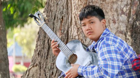 Téléchargez les photos : Jeune garçon asiatique joue de la guitare dans un parc local, accent doux et sélectif - en image libre de droit
