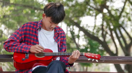 Téléchargez les photos : Jeune garçon asiatique joue de la guitare dans un parc local, accent doux et sélectif - en image libre de droit