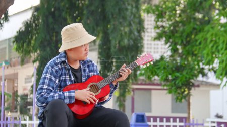 Foto de Joven chico asiático está tocando la guitarra en un parque local, enfoque suave y selectivo - Imagen libre de derechos