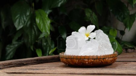A basket filled with white alum crystals sits on a vibrant green shrub background. The contrasting colors highlight the purity of the alum against the lush greenery, creating fresh feeling.