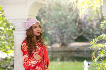 Téléchargez les photos : Une belle femme marocaine porte une robe marocaine traditionnelle en rouge et brodée d'or et d'argent, sur sa tête une écharpe de danse du ventre rose. Elle s'appuie sur une colonne de marbre. - en image libre de droit