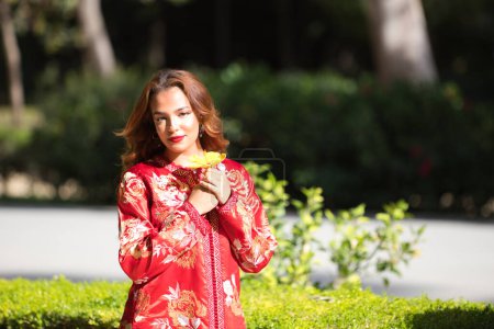 Téléchargez les photos : Une belle femme porte une robe traditionnelle marocaine en rouge et brodée d'or et d'argent. La fille tient une fleur jaune dans ses mains et la tient près de son visage. - en image libre de droit