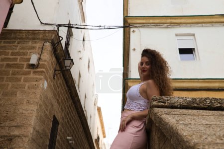 Foto de Hermosa mujer rubia con el pelo rizado y ojos azules apoyados en la pared de la escalera que sube al barrio de la ciudad donde está de vacaciones. Concepto de viaje. - Imagen libre de derechos