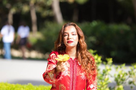 Téléchargez les photos : Une belle femme porte une robe traditionnelle marocaine en rouge et brodée d'or et d'argent. La fille tient une fleur jaune dans ses mains et la montre à la caméra. - en image libre de droit