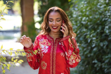 Téléchargez les photos : Une belle femme porte une robe traditionnelle marocaine en rouge et brodée d'or et d'argent. La belle femme parle au téléphone très heureux. - en image libre de droit