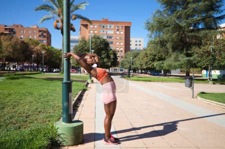 Téléchargez les photos : Jeune et belle Afro-Américaine au corps sculpté s'étirant dans le parc avant l'entraînement. Elle porte un haut orange et des leggings orange. Concept de sport et de santé. - en image libre de droit