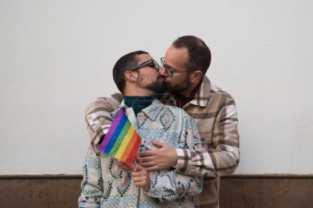 Ein junges Paar homosexueller Männer. Die Ehe ist glücklich und sie umarmen und küssen sich mit der Gay Pride Flagge in den Händen. Konzept für die Rechte Homosexueller.