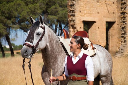 Photo for Young and beautiful Spanish woman with her thoroughbred horse. The woman is wearing a typical Spanish riding uniform. They are next to the stable in the countryside in seville. Concept horses. - Royalty Free Image