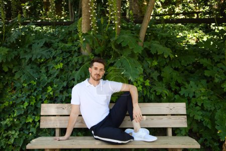 Photo for Handsome young man in white T-shirt and black trousers sitting on the wooden bench in the park waiting impatiently for someone to arrive. The man makes different body expressions. Concept expression. - Royalty Free Image