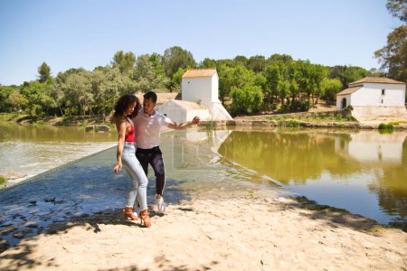Foto de Beautiful woman and handsome man latinos dancing bachata are dancing by the river in the forest. The couple do different postures while dancing. Dancing concept and expressions. - Imagen libre de derechos