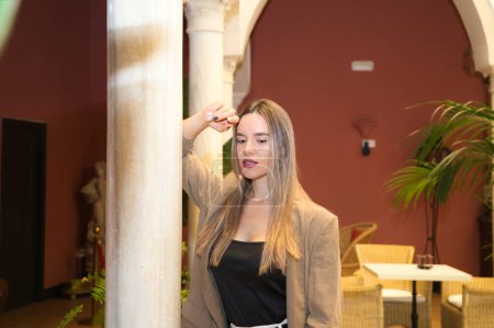 Foto de Young and beautiful blonde Latin woman is leaning against a marble column in the inner courtyard of a hotel. The woman is sad and pensive with anguish. Concept depression and stress. - Imagen libre de derechos