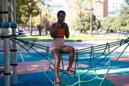 Photo for Young and beautiful Afro American woman with sculpted body sitting in the park before training. She is dressed in an orange top and tights and listens to music with white headphones. Sport and health. - Royalty Free Image
